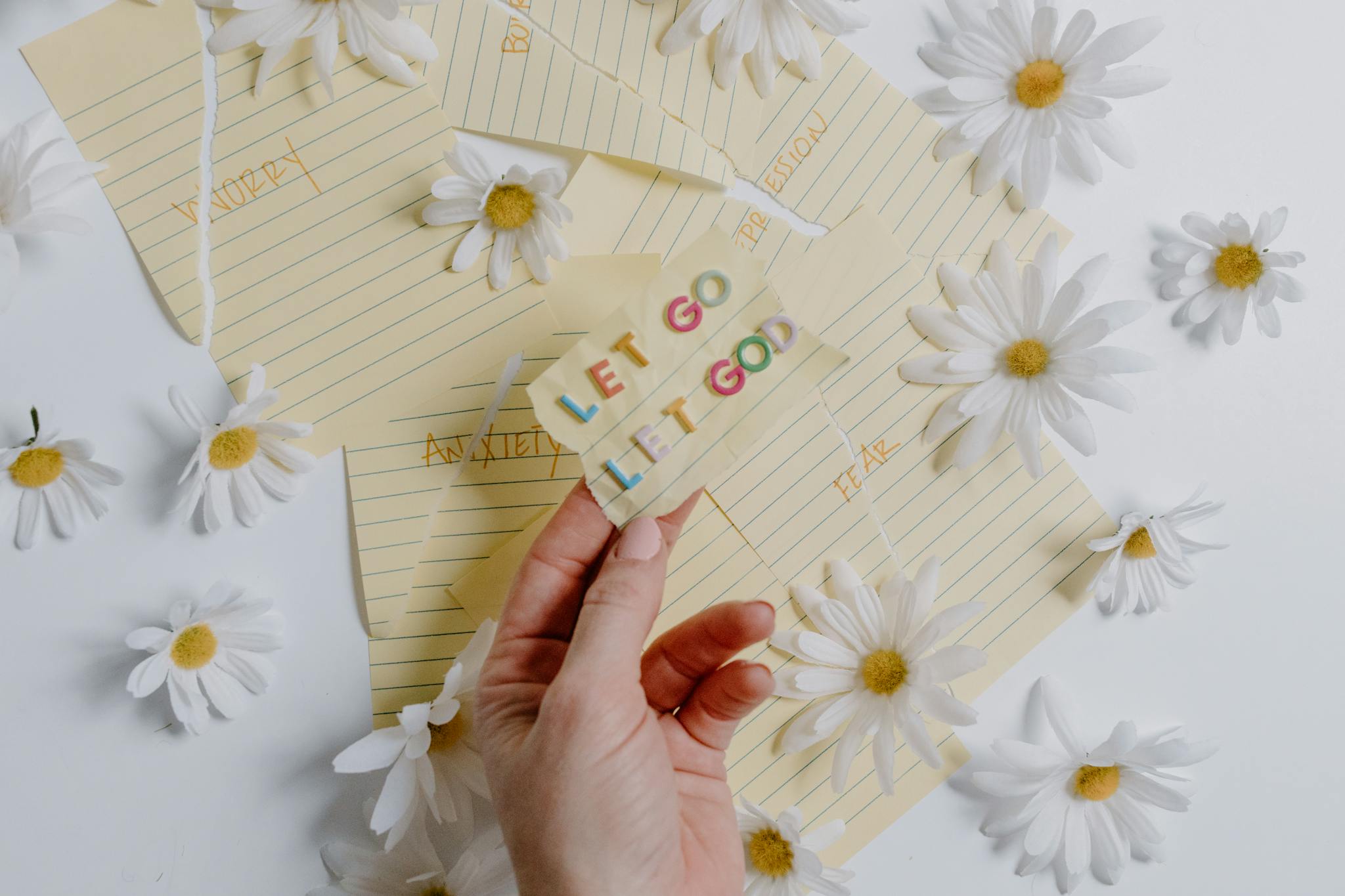 Hand holding a note with 'Let Go, Let God' surrounded by daisies on lined paper.